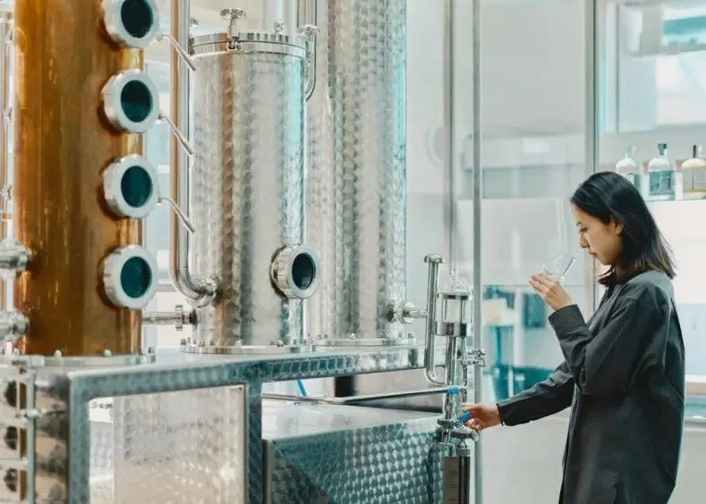 蒸留所のような施設で、女性が大型の機械の横でグラスに入った液体をテイスティングしている様子