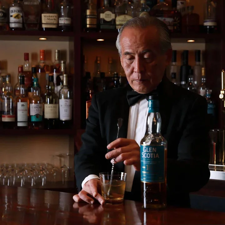 タキシードを着たベテランのバーテンダーが、バーカウンターでグラスに入ったウイスキーを丁寧に混ぜています。背景には多くの酒瓶が並び、手前には「GLEN SCOTIA」のボトルが置かれています。
