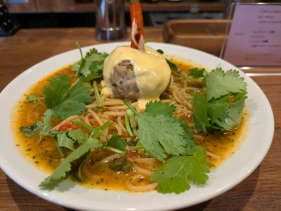 大皿に盛られたパスタ料理で、大きなミートボールに黄色いソースと赤唐辛子が添えられ、フレッシュなパクチーがふんだんに飾られている