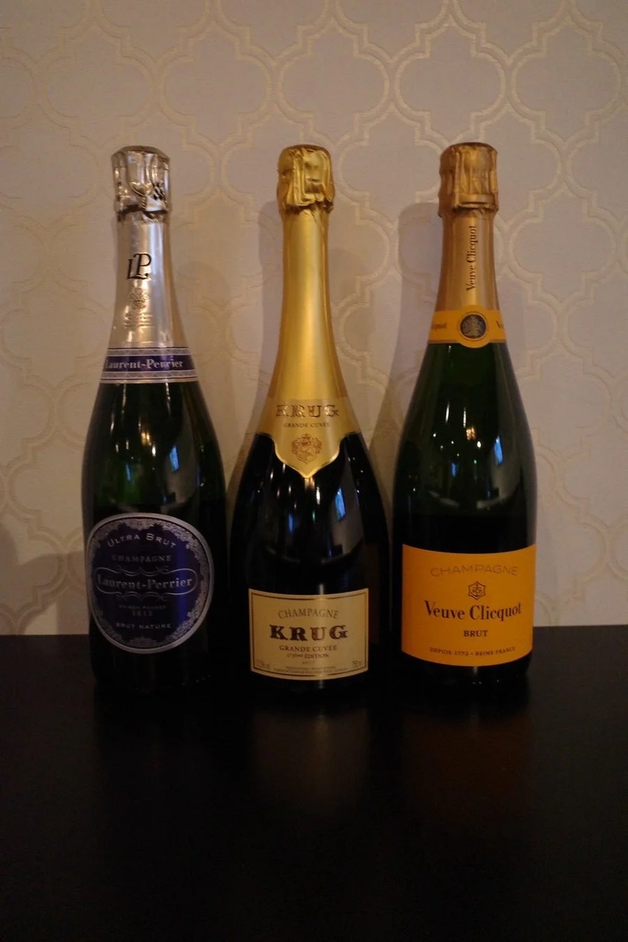 ローラン・ペリエ、クリュッグ、ヴーヴ・クリコといった3種類のシャンパンボトルが、暗いテーブルと模様入りの壁を背景に並べられています。