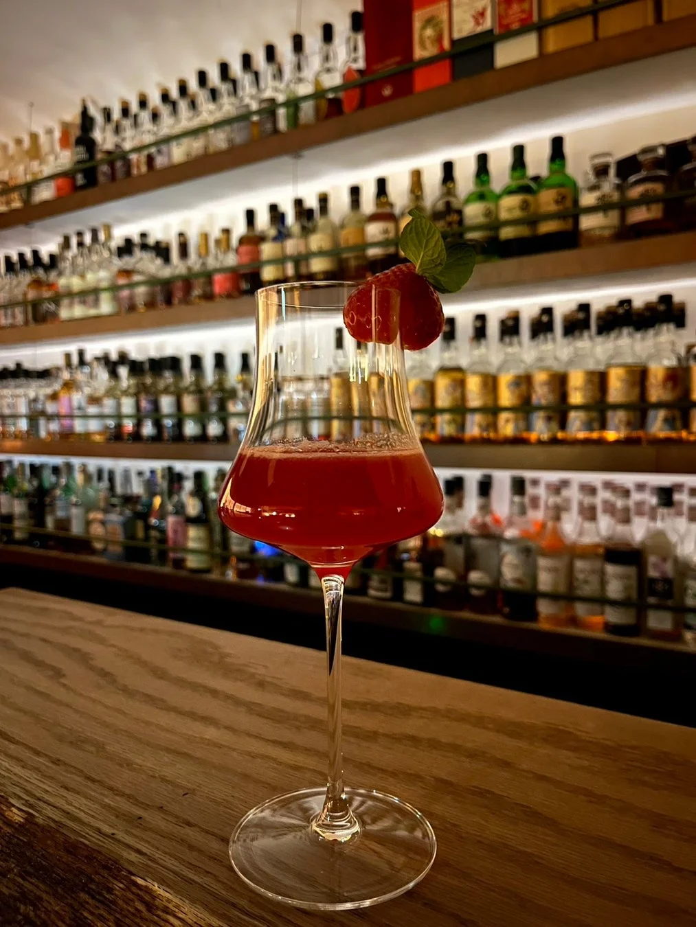 バーの木製カウンターに置かれた、イチゴとミントの葉で飾られた赤いカクテルです。背景にはライトアップされた棚に多くの酒瓶が並び、落ち着いた雰囲気を示しています。