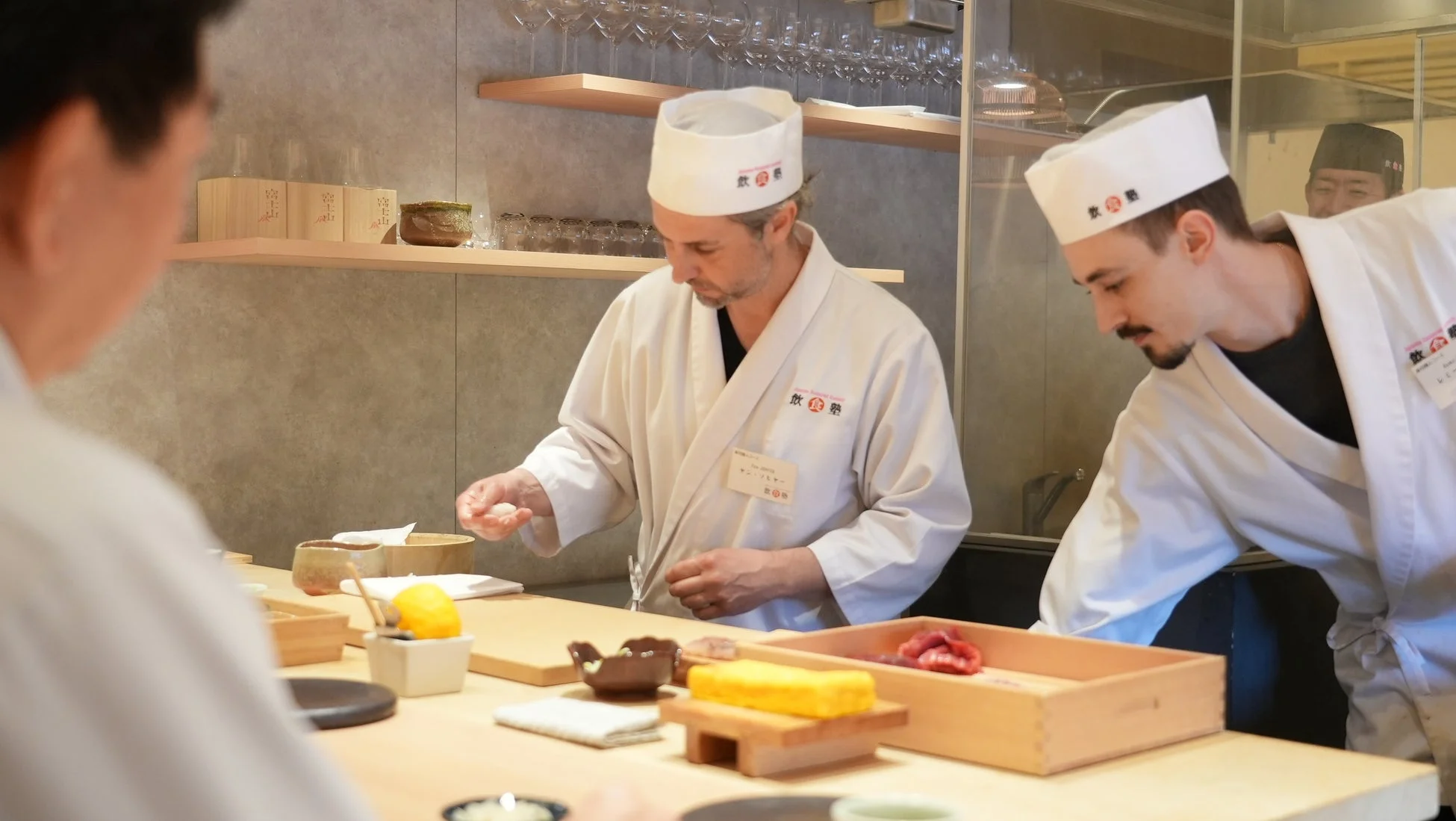 和食レストランのカウンターで、白衣を着た2人の寿司職人が寿司を準備している様子。手元には握り飯や食材が見えます。