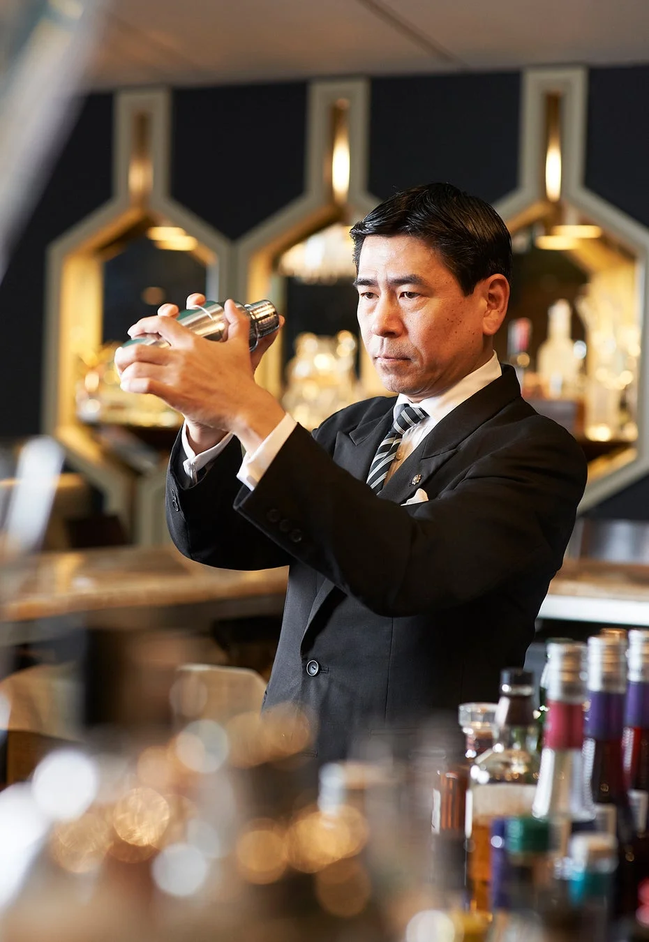 バーカウンターで黒いスーツの男性バーテンダーが、集中した表情でシェイカーを振ってカクテルを調合しています。高級感のあるバーの雰囲気が伝わる一枚です。