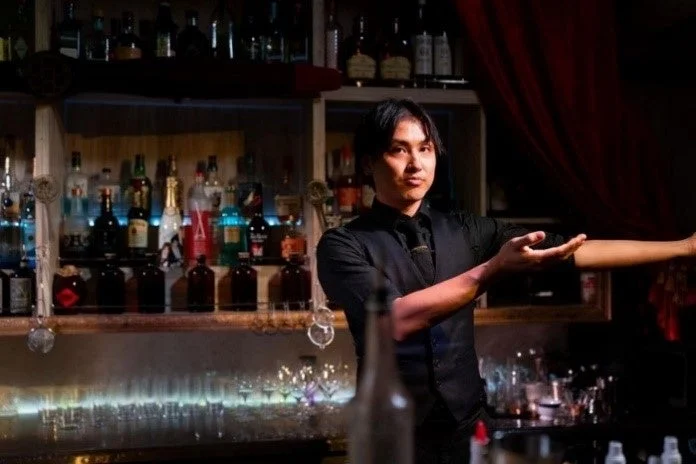 黒いベストとシャツを着た男性バーテンダーが、豊富な酒瓶とグラスが並ぶバーカウンターの前に立ち、手で何かを案内しているようなポーズをとっています。