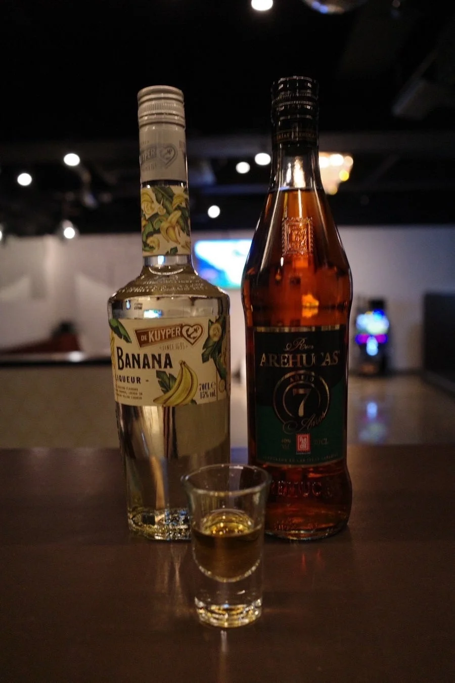 暗いバーのような空間に、バナナリキュールと7年熟成のラム酒のボトルが並び、手前にはショットグラスに注がれたお酒がある。ナイトライフを思わせる雰囲気。