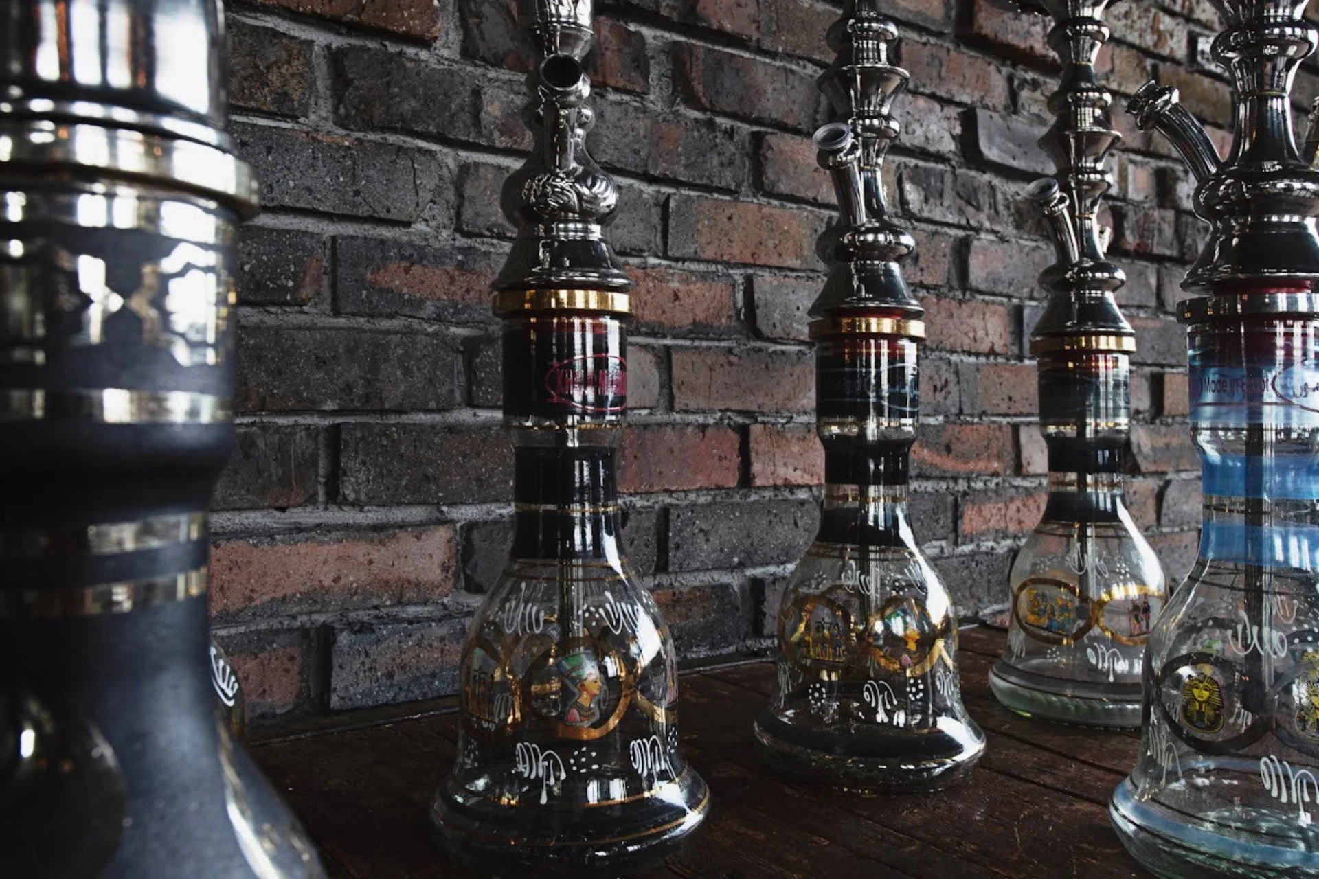 レンガの壁を背景に、精巧な装飾が施された複数の水タバコが並んでいます。ガラス製の本体にはエジプト風の模様が見られ、異国情緒あふれる雰囲気です。