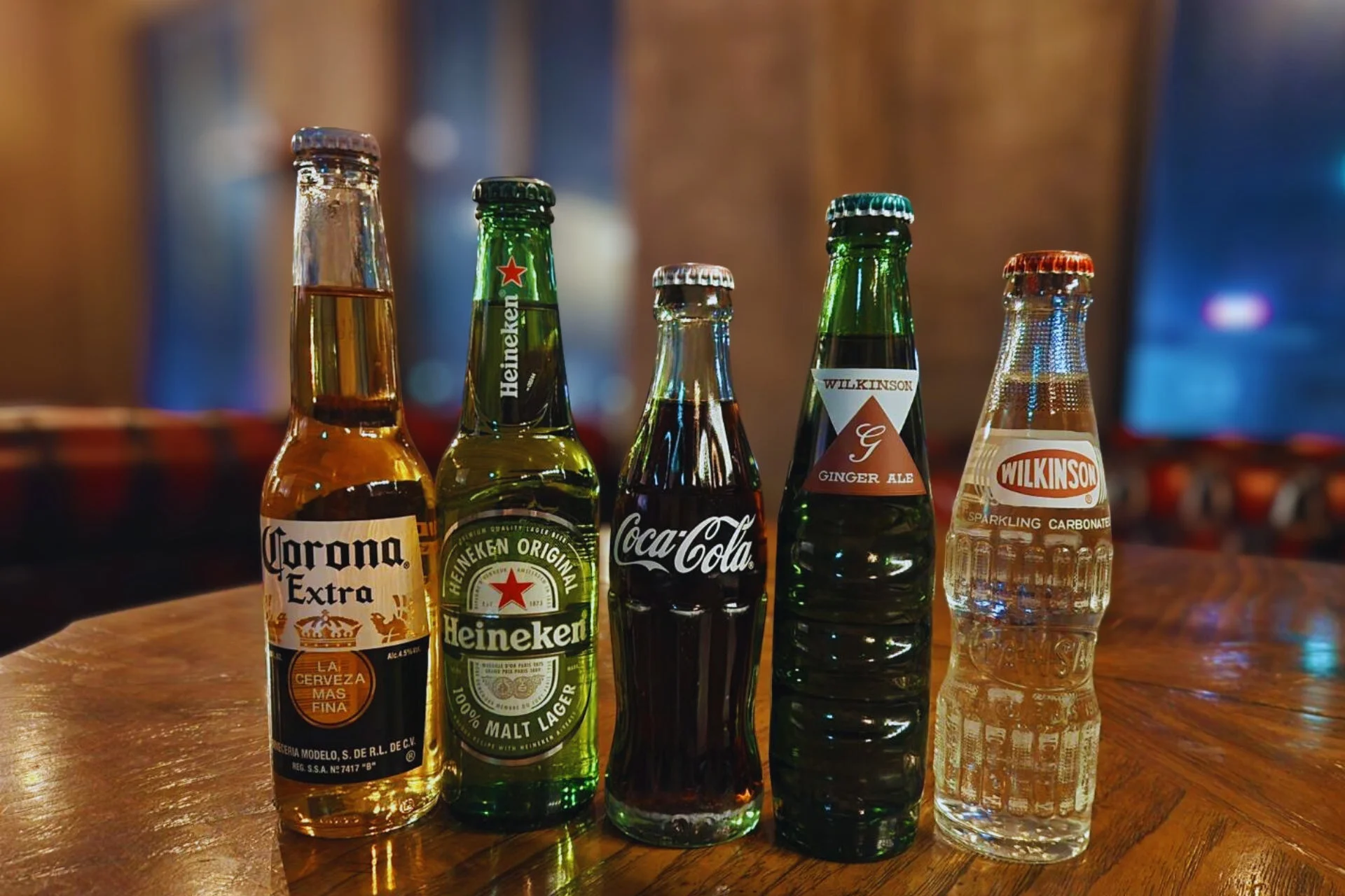 木製のテーブルの上に、コロナエクストラ、ハイネケン、コカ・コーラ、ウィルキンソンジンジャーエール、ウィルキンソン炭酸水といった様々な種類の瓶入り飲料が5本並べられています。背景はぼやけており、飲食店のような雰囲気が感じられます。