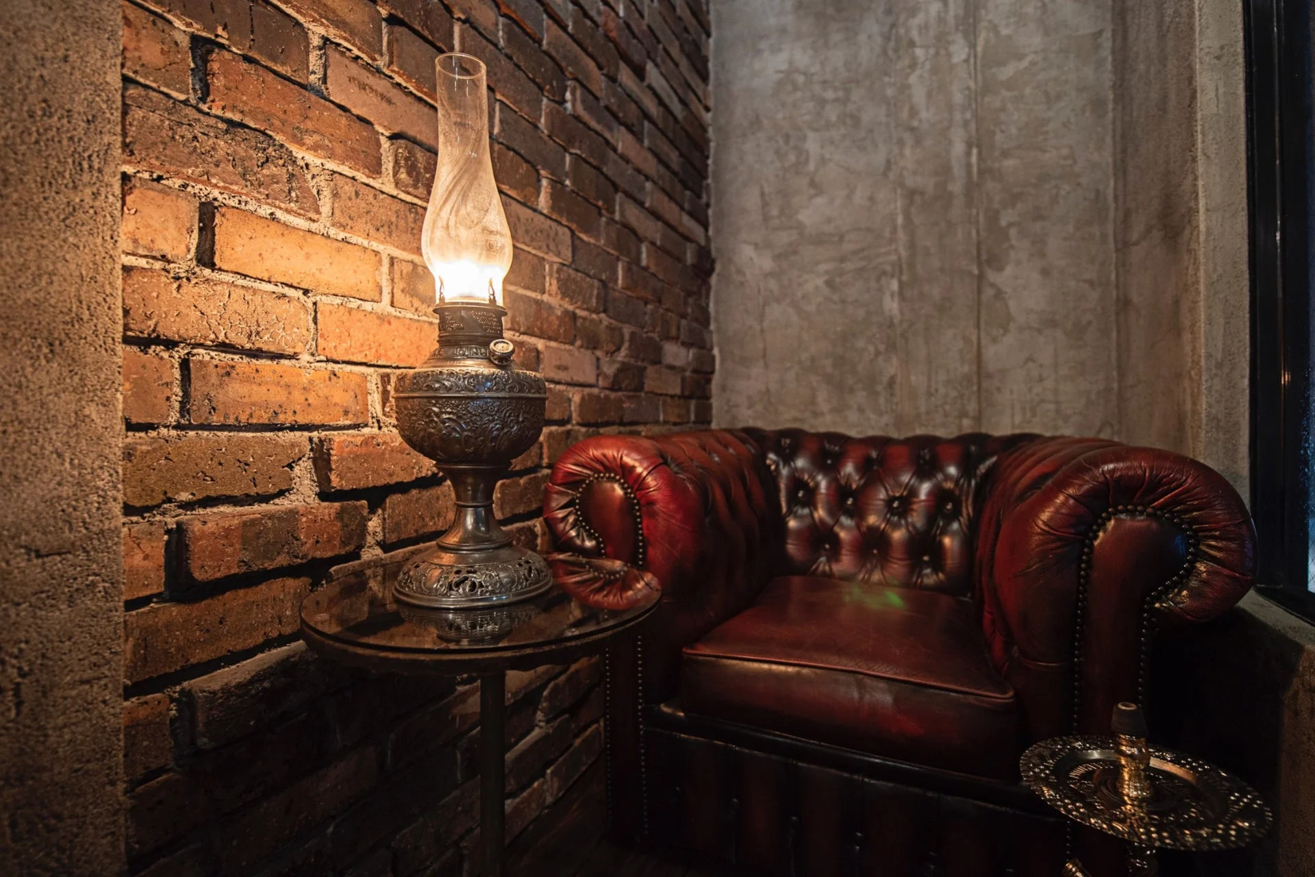 温かい光を放つアンティークなオイルランプが、レンガとコンクリートの壁に囲まれた空間を照らしています。隣には深紅の革製チェスターフィールドソファが置かれ、落ち着いた大人の雰囲気を醸し出しています。