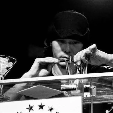 帽子をかぶった男性が、グラスや細い棒のようなものに集中しているモノクロ写真です。