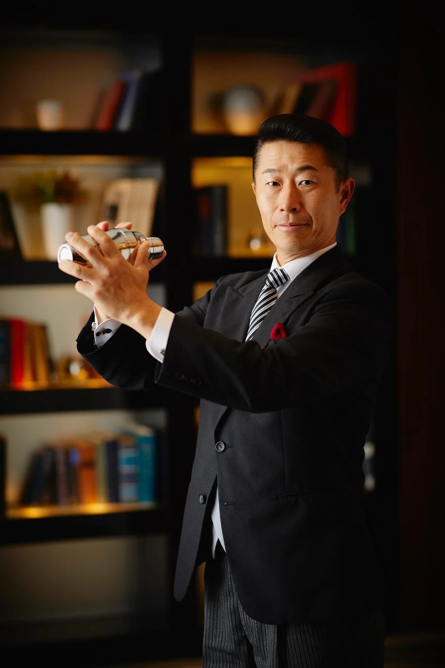 スーツを着た男性がカクテルシェイカーを両手で持ち、カメラを見ているポートレートです。高級感のあるバーカウンターのような場所で、プロのバーテンダーがカクテルを作っている様子を捉えています。