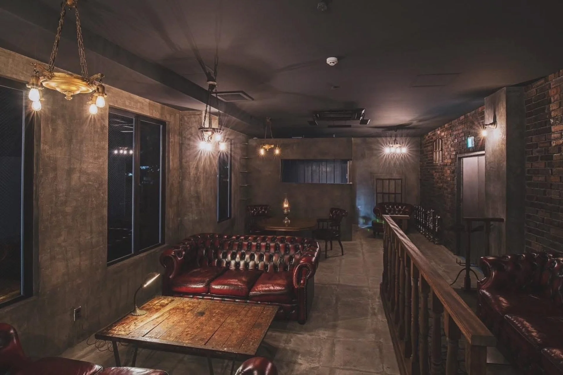 ヴィンテージ感のある落ち着いた雰囲気のラウンジ空間です。深紅のチェスターフィールドソファや木製のテーブルが配置され、コンクリートとレンガの壁がインダストリアルな魅力を醸し出しています。暖色の照明が心地よい、くつろぎの場所です。