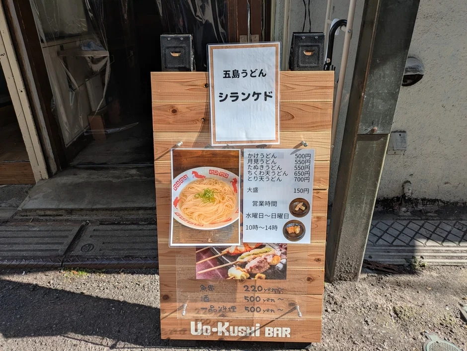 五島うどんを提供する「Uo-Kushi BAR」の店頭に設置された木製看板の画像です。看板には、かけうどんや月見うどんなどのうどんメニューと価格、営業時間（水～日 10時～14時）、そして魚や酒などのサイドメニューが詳細に表示されています。