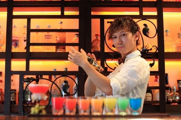 バーカウンターで男性バーテンダーがシェイカーを振っており、手前には虹色に並んだカラフルなカクテルが置かれています。