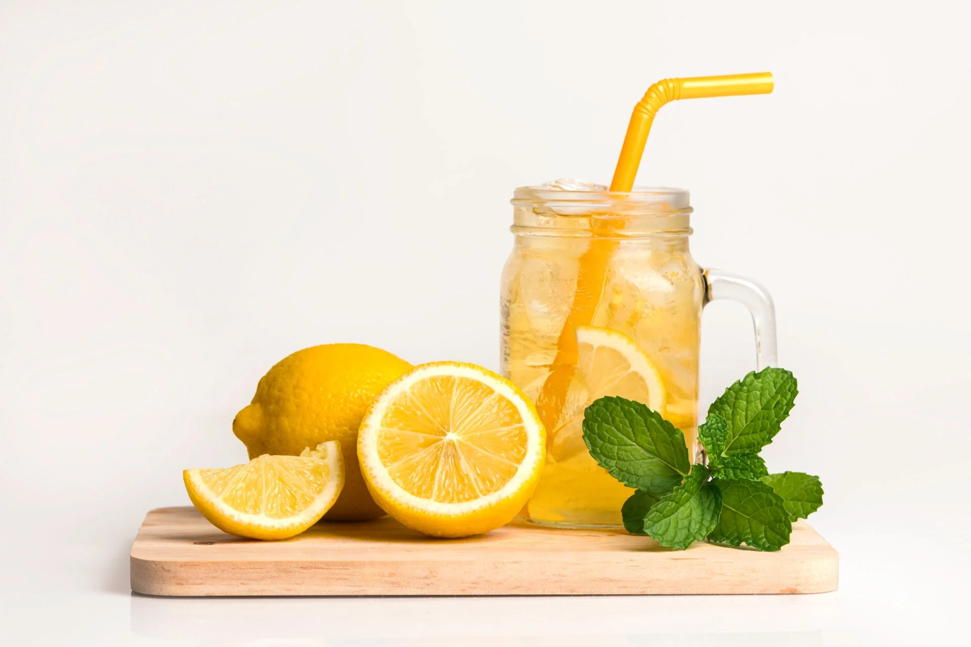 氷とレモンスライスが入ったレモネードのグラスと、新鮮なレモン、ミントの葉が木製のボードに置かれている。爽やかな夏の飲み物を連想させる写真です。
