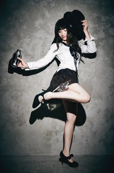 白いシャツと黒いスカートを着用した女性が、カクテルシェーカーと帽子を持ち、躍動的なポーズで立っている。