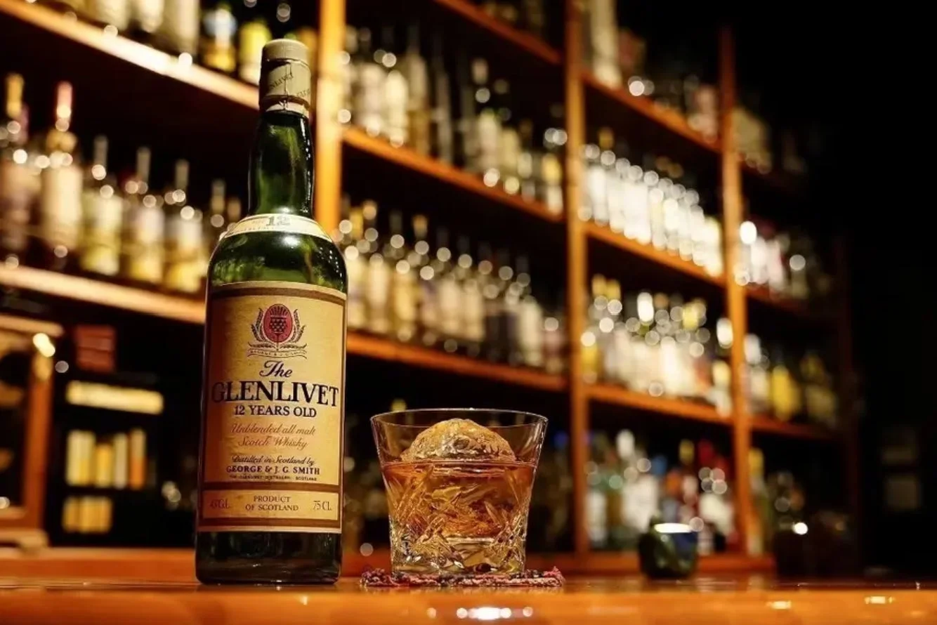 バーのカウンターに置かれたグレンリベット12年のボトルと、大きな丸氷が入ったロックグラスのウイスキーが写っています。背景には多くの酒瓶が並ぶ棚があり、落ち着いた大人の雰囲気を醸し出しています。