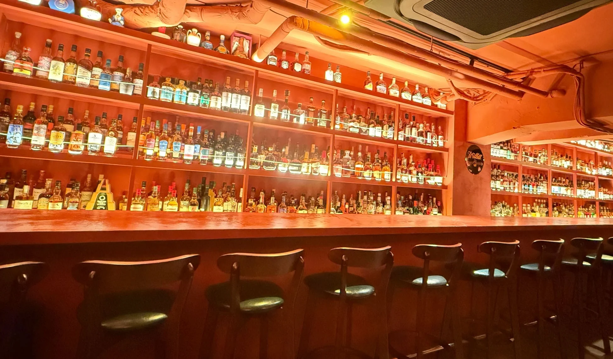多数の酒瓶が並ぶ棚とカウンター、バーチェアが特徴的なバーの店内。オレンジ色の温かい照明が落ち着いた雰囲気を醸し出している。