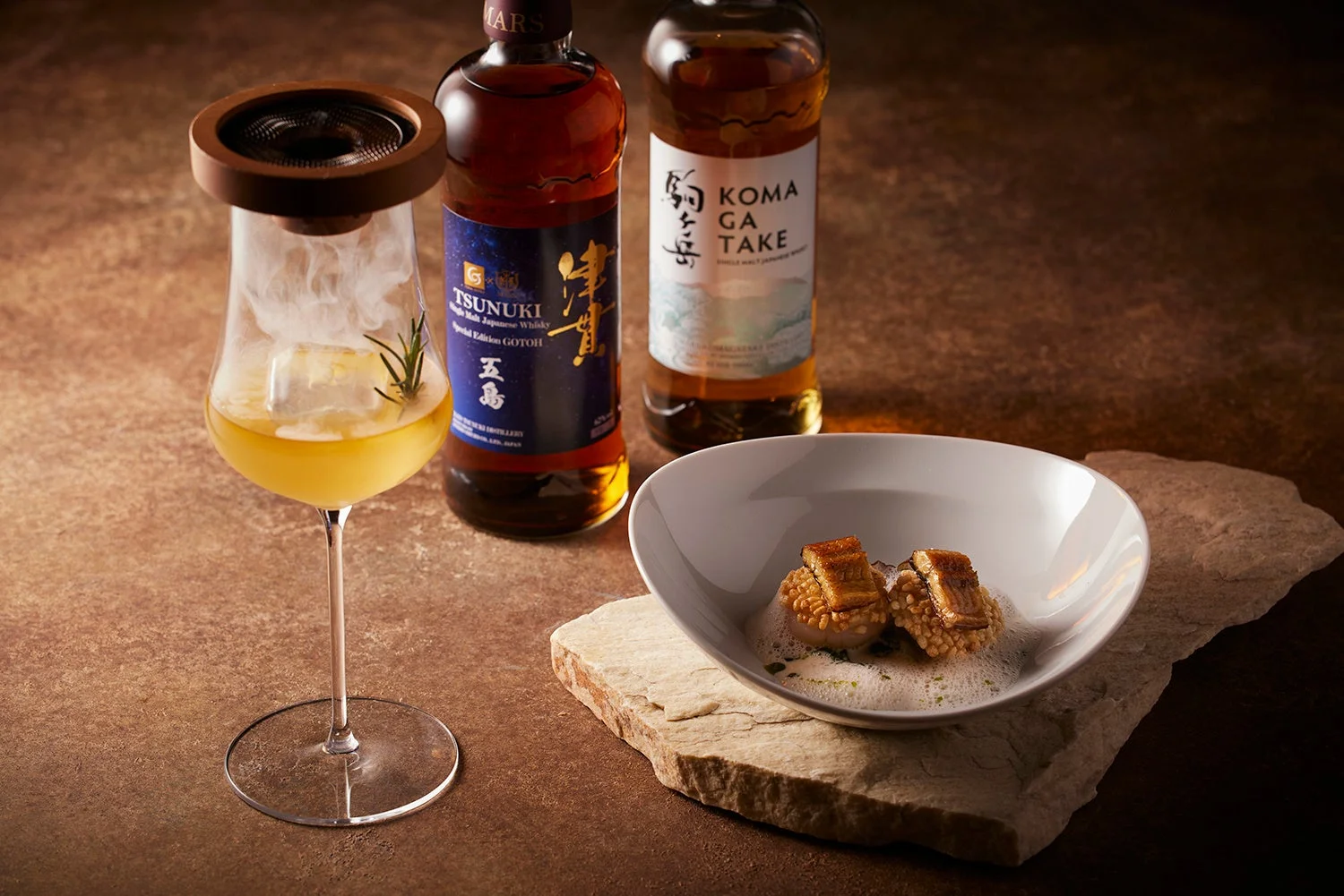 煙をまとったウイスキーカクテル、日本のシングルモルトウイスキー2本（ツヌキ、駒ヶ岳）、そして器に盛られた高級感のある料理が写っています。ウイスキーと料理のペアリングを思わせる、洗練された雰囲気の画像です。