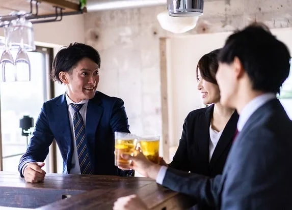 ビジネスウェアを着た男女3人が、バーカウンターでビールグラスを手に笑顔で乾杯している様子