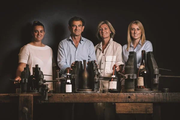 ワインボトルと古い機械が並ぶ作業場で、4人の男女が微笑んでいます。ワイン製造に関わる家族またはチームのポートレートです。
