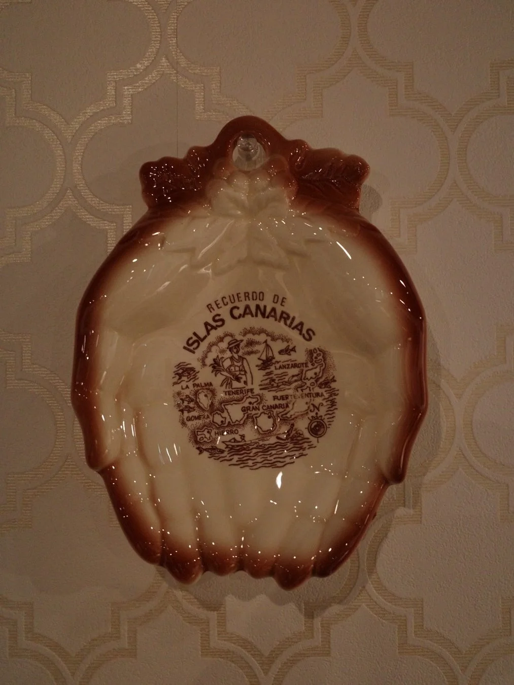 壁に飾られた、貝殻の形をしたカナリア諸島のお土産の陶器の皿です。皿の中央にはカナリア諸島の地図と「RECUERDO DE ISLAS CANARIAS」（カナリア諸島の思い出）という文字が描かれています。