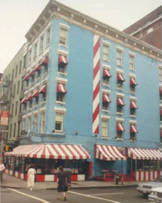 青い壁の建物が特徴的な街角の風景。建物には赤と白のストライプの日よけと、同じデザインの屋根を持つ店舗があり、人々が通りを歩いている。