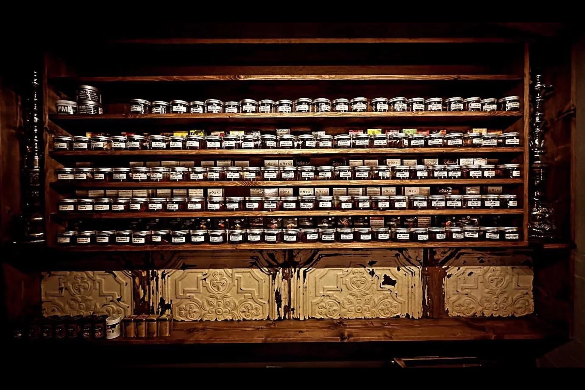 暗い照明の部屋にある木製の棚に、ラベルの貼られた小さな瓶や缶が多数整然と並べられています。DOZAJと書かれた瓶が多く、スパイスや食品などのコレクションが美しく陳列されている様子です。