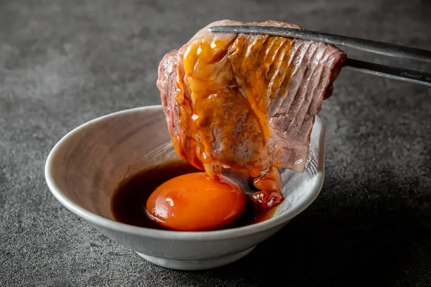 箸で掴んだ焼肉を、醤油ベースのたれと生卵の卵黄が入った小鉢に浸している