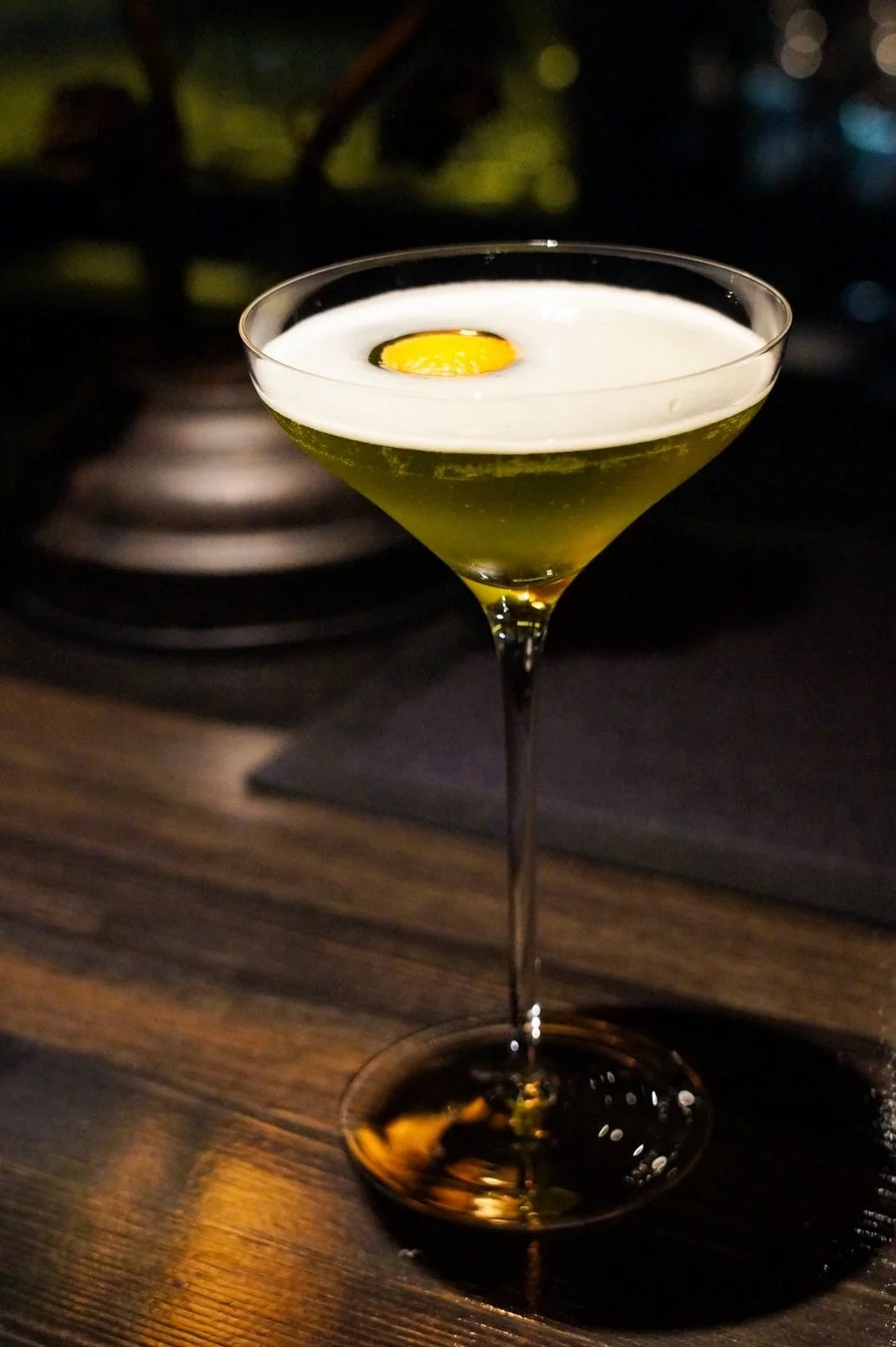 暗いバーのカウンターに置かれた、白い泡と黄色のガーニッシュが特徴的な緑がかったカクテル