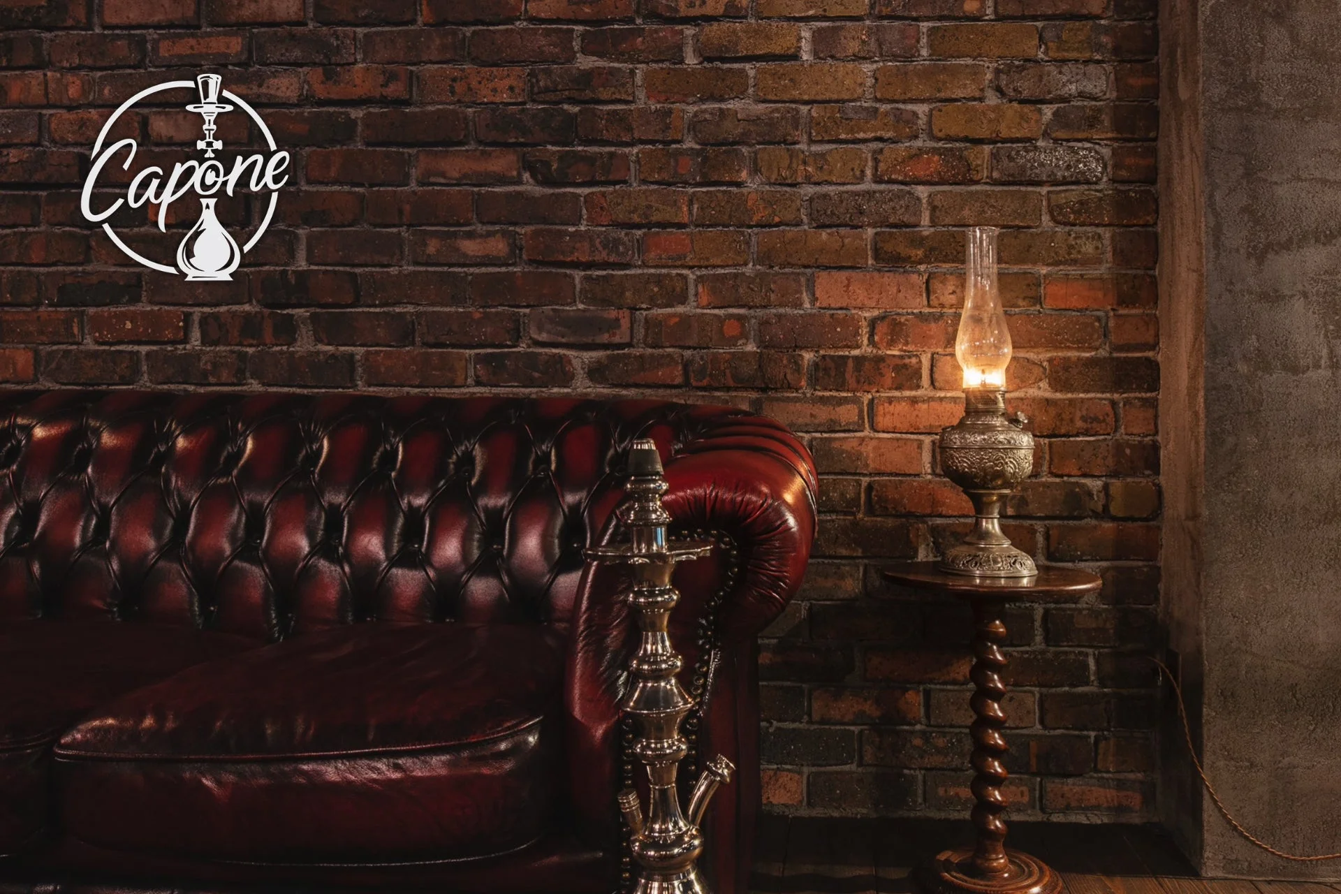 赤色の革製チェスターフィールドソファが置かれた、レンガ壁の室内空間です。手前にはシーシャ、奥にはアンティーク調のランプが灯るサイドテーブルがあり、落ち着いたレトロな雰囲気を醸し出しています。左上には「Capone」と書かれたロゴが見えます。