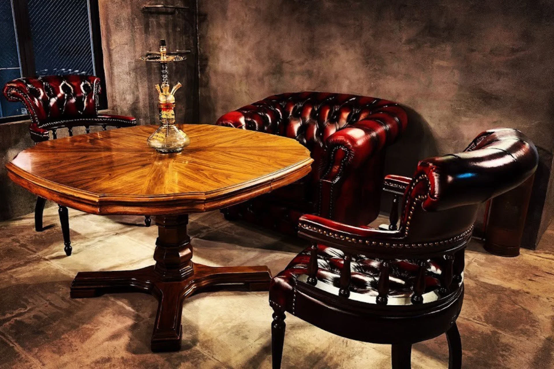 深紅のチェスターフィールドソファと椅子が並ぶ、落ち着いた雰囲気の室内。木製テーブルにはシーシャが置かれ、ヴィンテージ感のある高級ラウンジやバーのような空間を演出している。