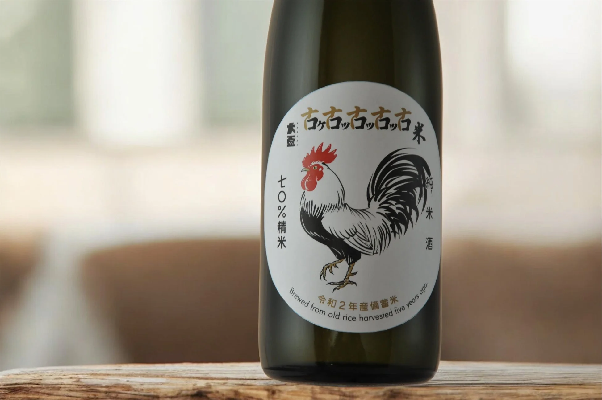 鶏のイラストが描かれた日本酒のボトルが木製のテーブルに置かれています。「古コッココッコ古米 純米酒」とあり、令和2年産の備蓄米を5年前に収穫された古米から醸造した純米酒であることが示されています。