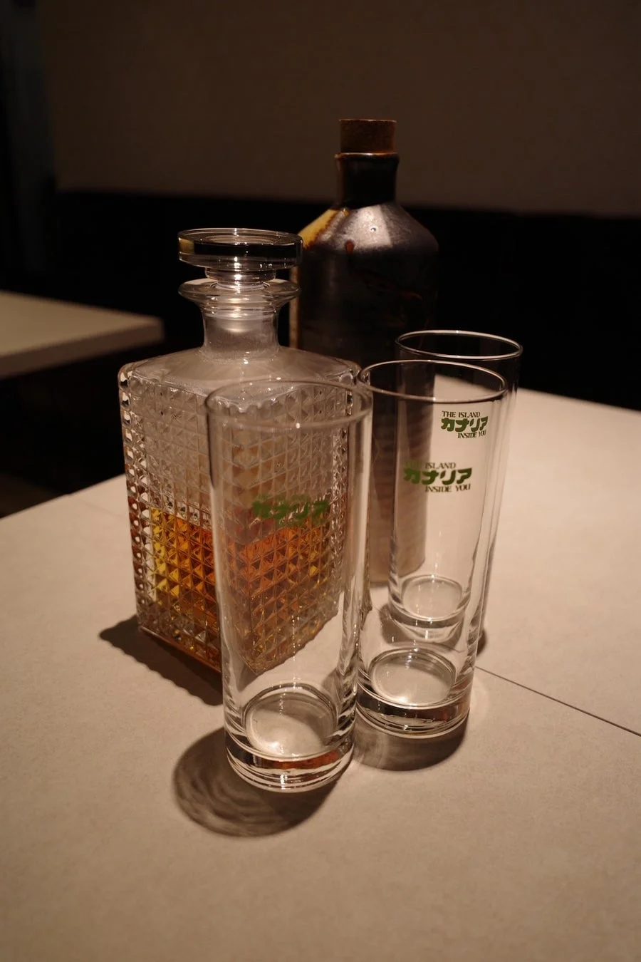薄暗い照明の下、テーブルに置かれたウイスキーデカンタ、陶器のボトル、そして2つのグラスが写っています。デカンタには琥珀色の液体が入っており、グラスには「THE ISLAND カプリラ INSIDE YOU」という文字が見えます。落ち着いた雰囲気で、バーや自宅での晩酌シーンを思わせる一枚です。