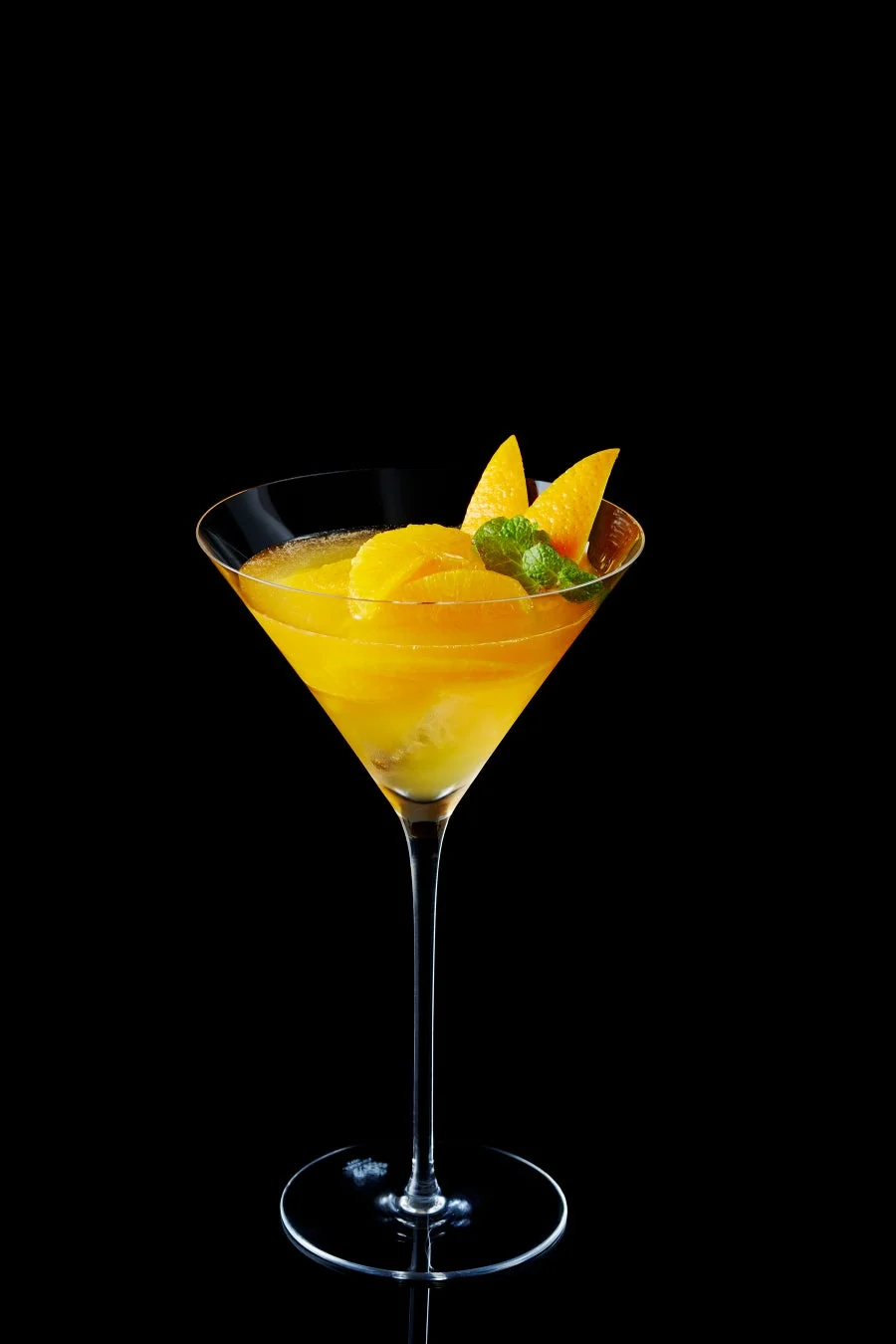 黒い背景にマティーニグラスが置かれ、鮮やかな黄色のドリンクが入っています。ドリンクはマンゴーやオレンジのスライス、ミントの葉で飾られており、夏らしい爽やかなカクテルやデザートを思わせる一枚です。