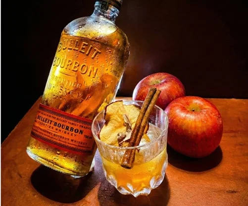 ブレットバーボンのボトルと、ドライアップルとシナモンスティックで飾られたバーボンベースのドリンク