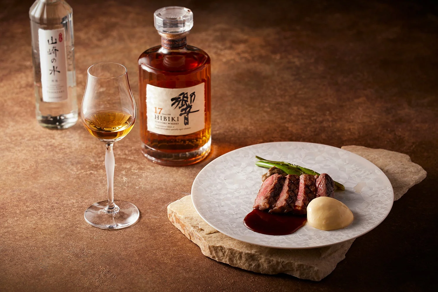 スライスされたステーキとウイスキー、そしてミネラルウォーターが並べられた、上質なディナーの様子です。響17年ウイスキーがグラスに注がれ、贅沢な食卓を演出しています。