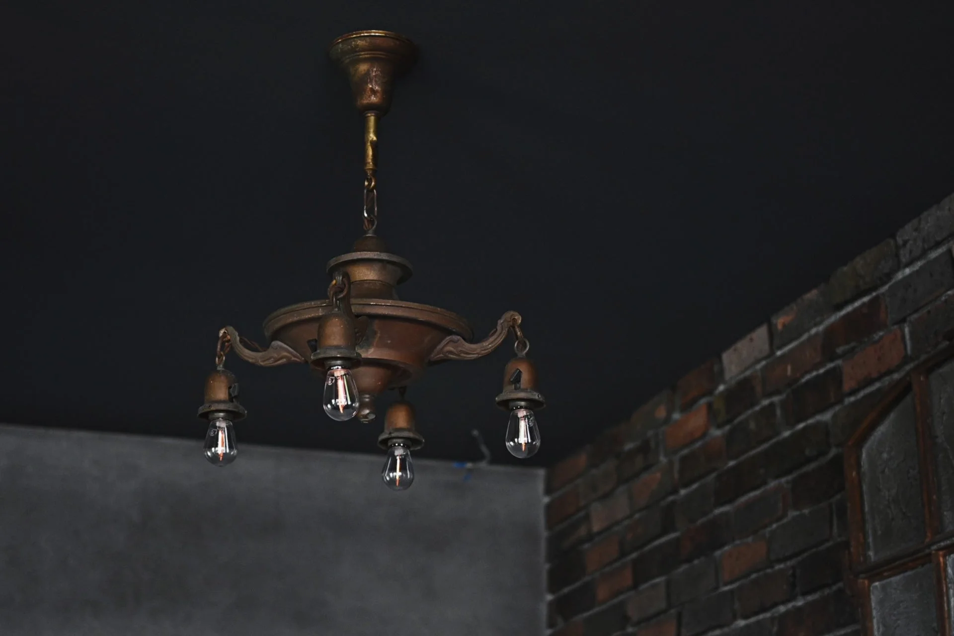 暗い天井から吊り下げられたレトロなシャンデリアが写っています。剥き出しの電球が特徴的で、右側にはレンガの壁が見えます。