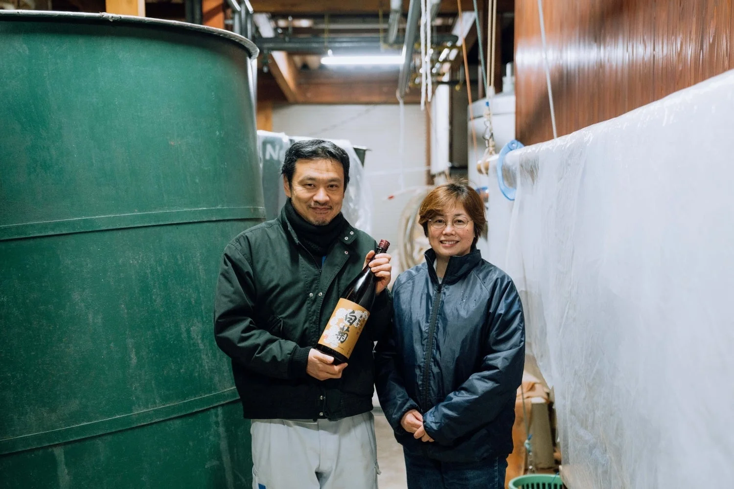 酒蔵のような場所で、笑顔の男女が日本酒のボトルを持って立っています