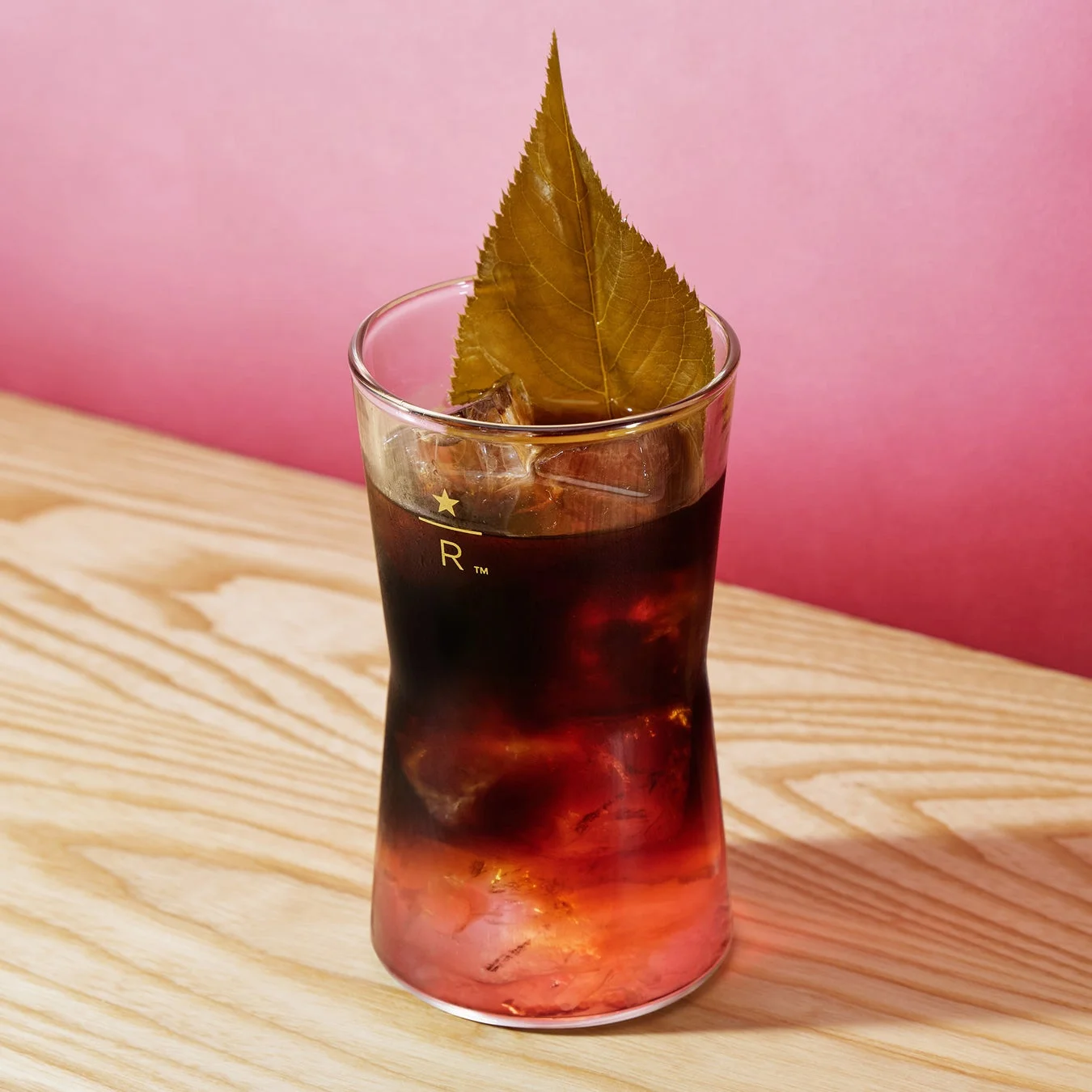 木製のテーブルに置かれた、ピンクの背景を背にしたグラス入りのアイスドリンクです。ドリンクは上部が濃い色で下部が赤みがかったグラデーションになっており、大きな葉っぱが飾られています。グラスには「R™」と星のロゴが見えます。