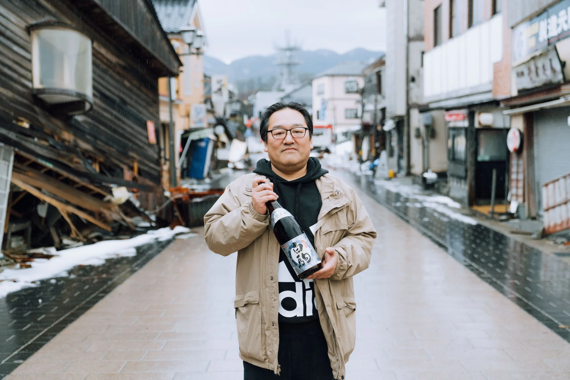 日本の雪が残る街並みで、眼鏡をかけた男性が日本酒のボトルを抱えて立っています