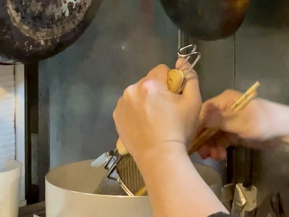 キッチンのような場所で、人物が両手でおろし器と箸を持ち、何かをすりおろしている瞬間