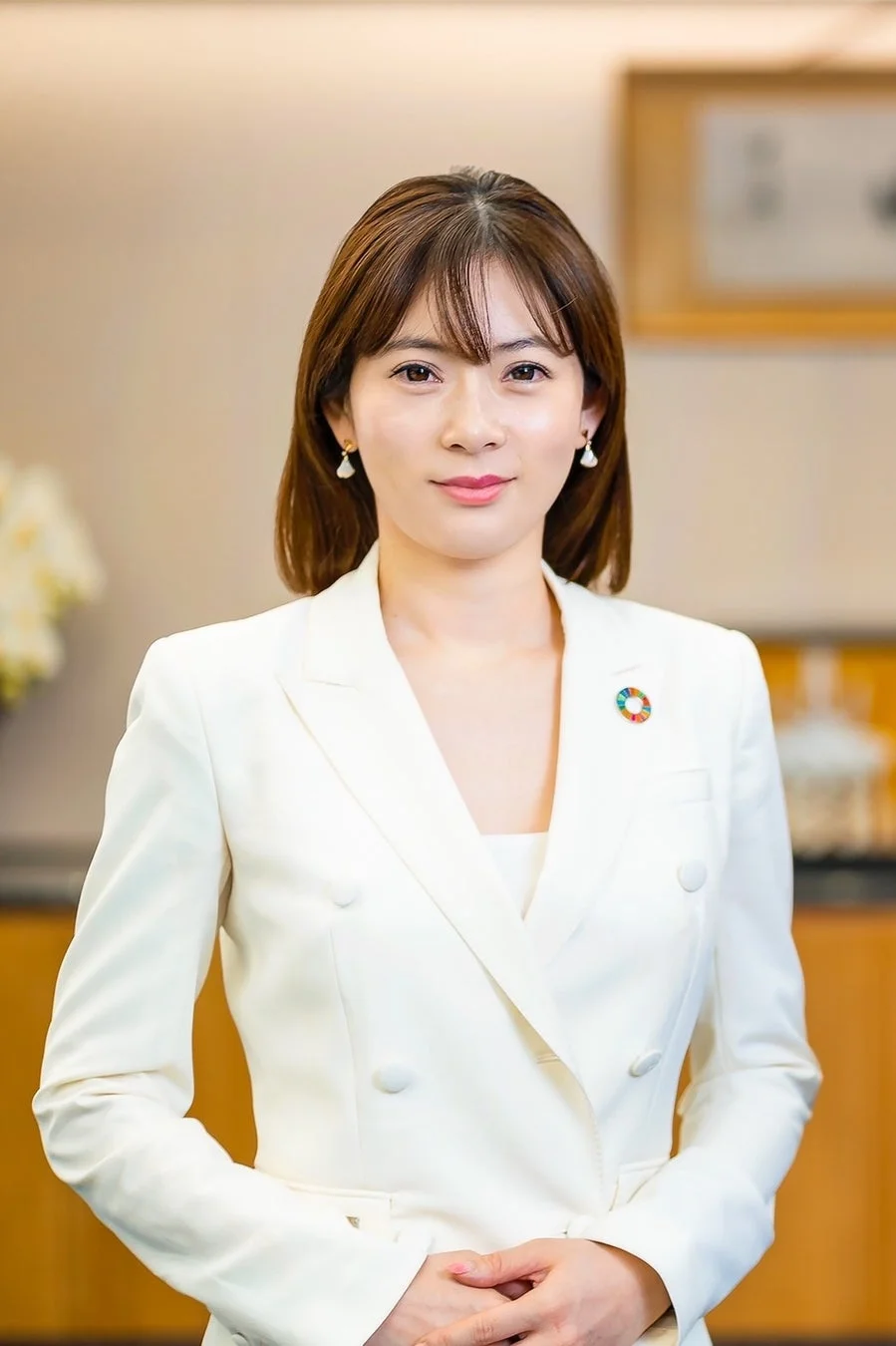 白いスーツを着用した女性が正面を向いて立ち、穏やかな笑顔を見せています。胸元にはSDGsのピンバッジが付けられており、プロフェッショナルで清潔感のある印象です。