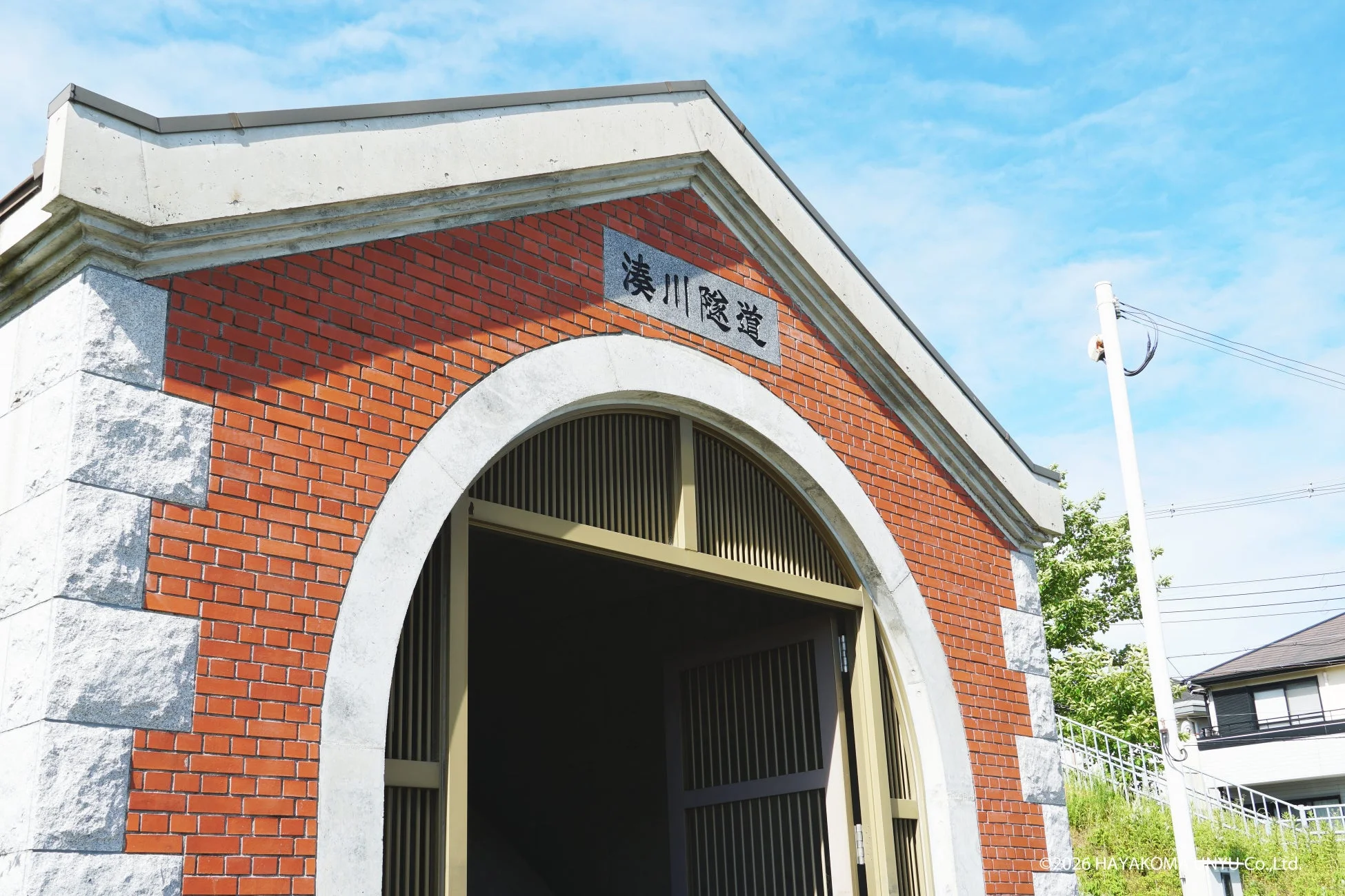 青空の下に立つ、赤レンガと石でできたレトロな建築物「湊川隧道」の入り口部分を捉えた画像です。