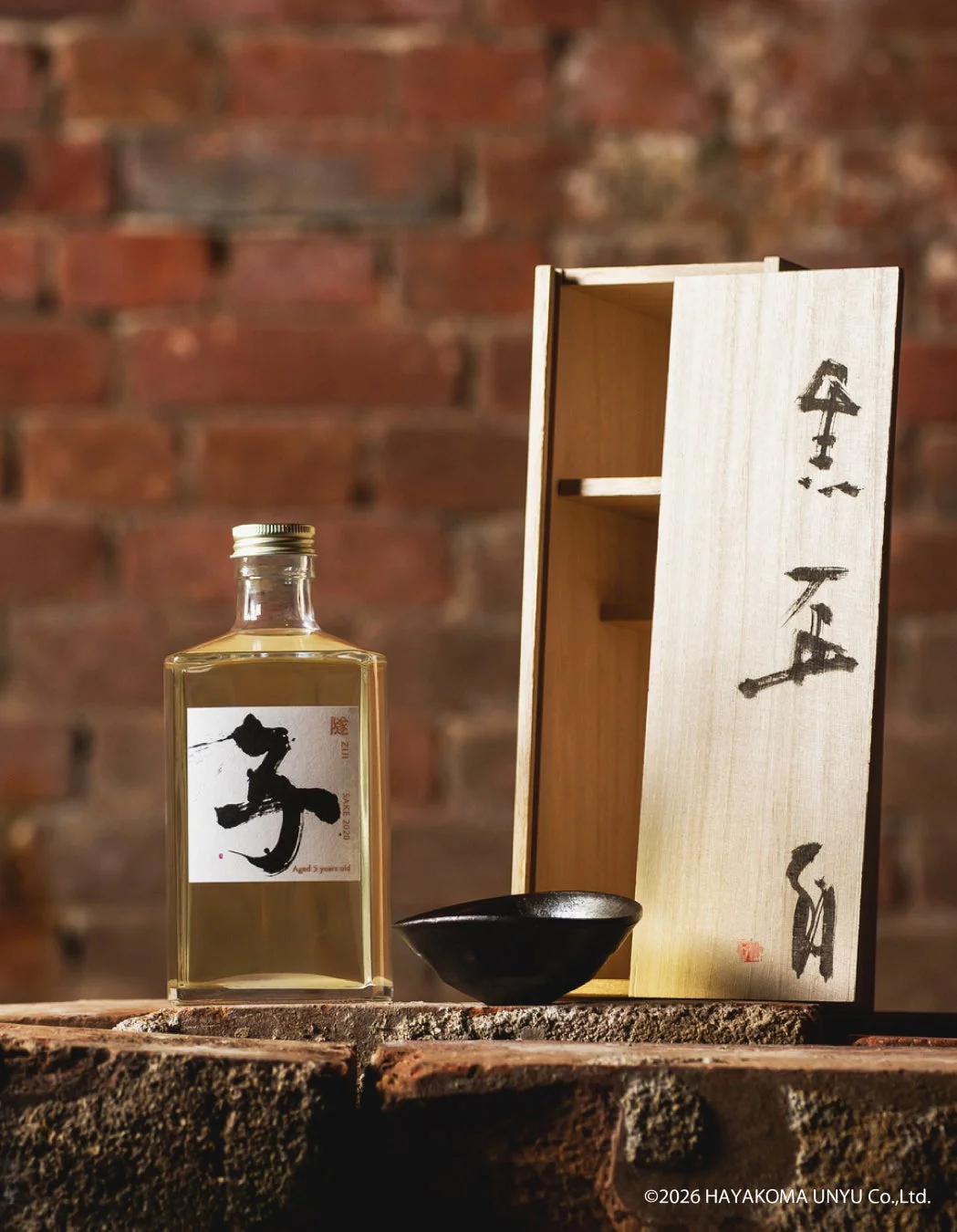 レンガ壁を背景に、熟成された日本酒「ZUI SAKE 2020 Aged 3 years old」のボトルと、書道が施された木箱、そして黒い陶器の盃が並べられています。