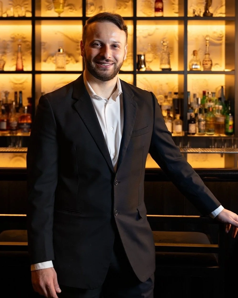 黒いスーツと白いシャツを着用した男性が、照明付きのバーカウンターを背景に笑顔で立つ