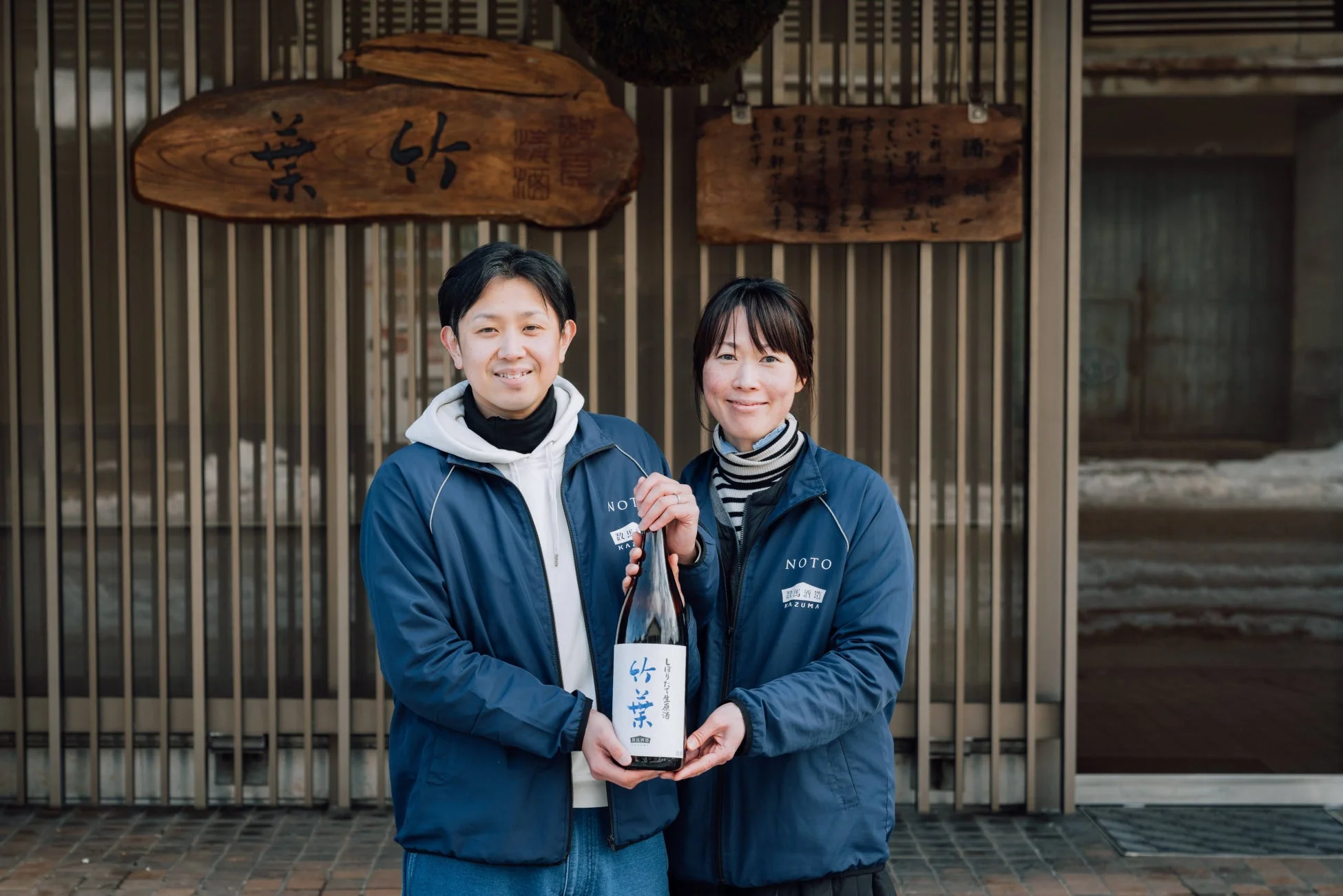 笑顔の男女が日本酒「竹泉」のボトルを持ち、伝統的な酒蔵の前でポーズをとっています