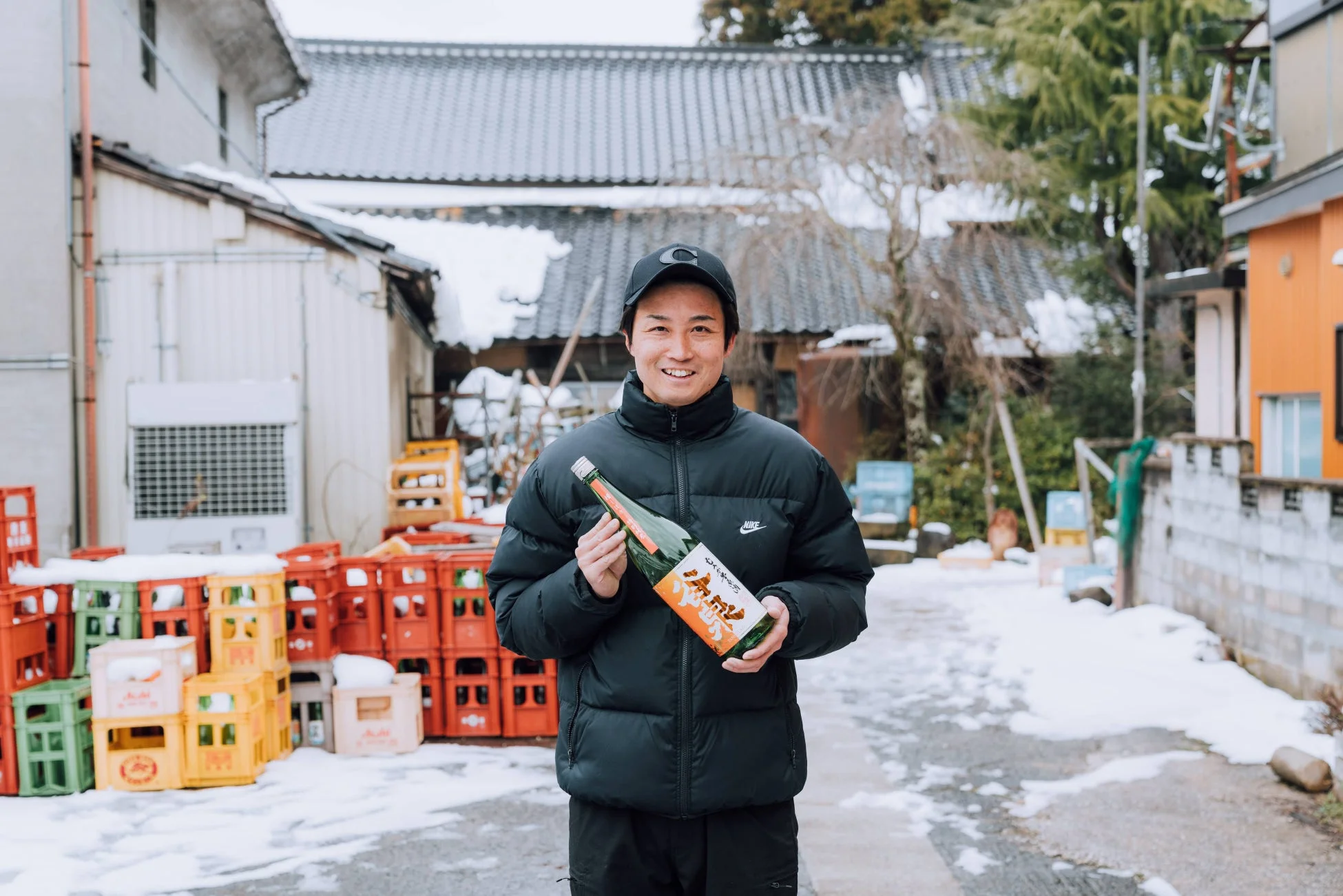 雪が積もる屋外で、黒いダウンジャケットとキャップを身につけた笑顔の男性が日本酒の瓶を両手に持っています