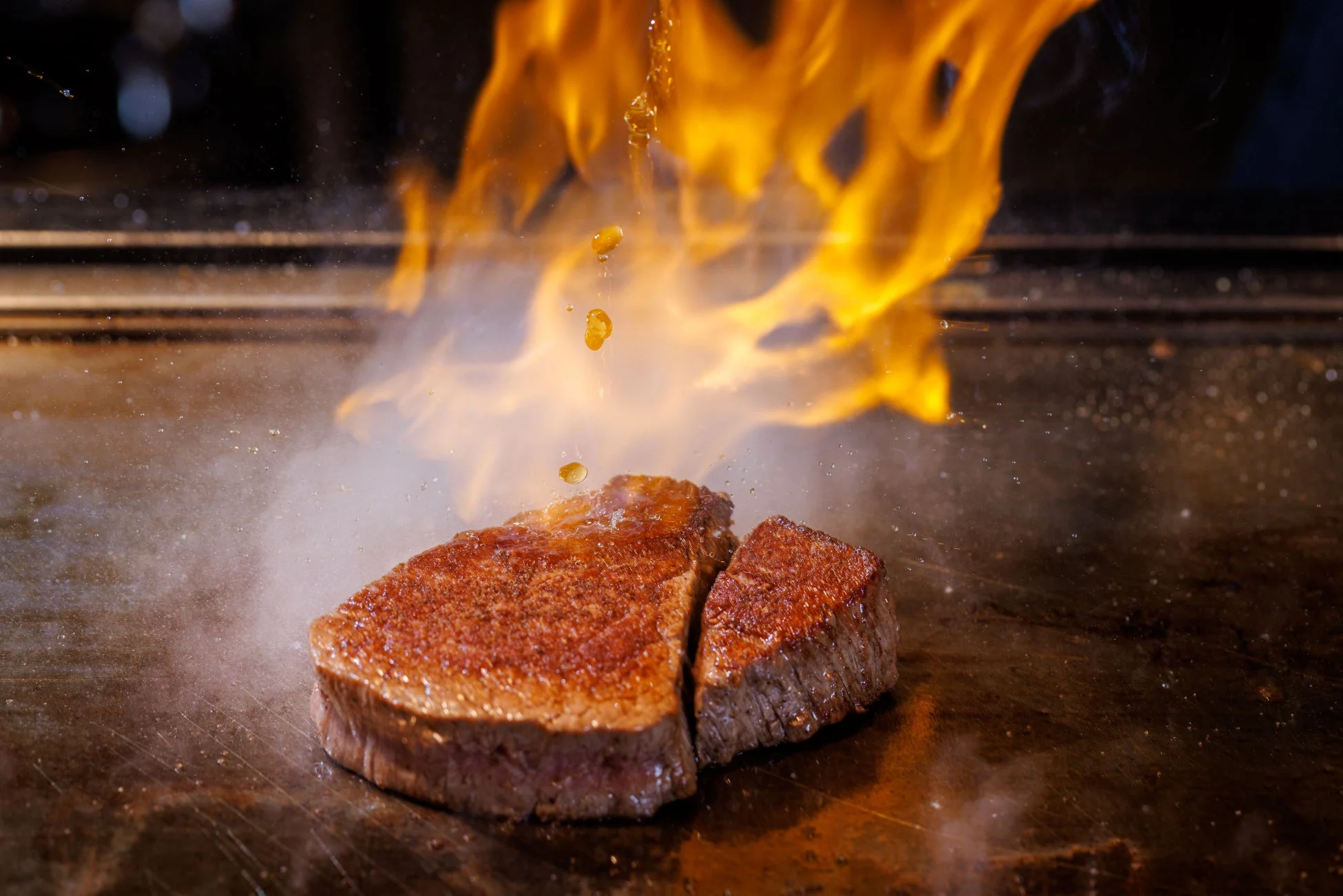 鉄板の上で分厚いステーキが炎を上げて調理されている瞬間を捉えた写真です。肉は香ばしく焼かれ、液体が滴り落ちる様子が臨場感を高めています。