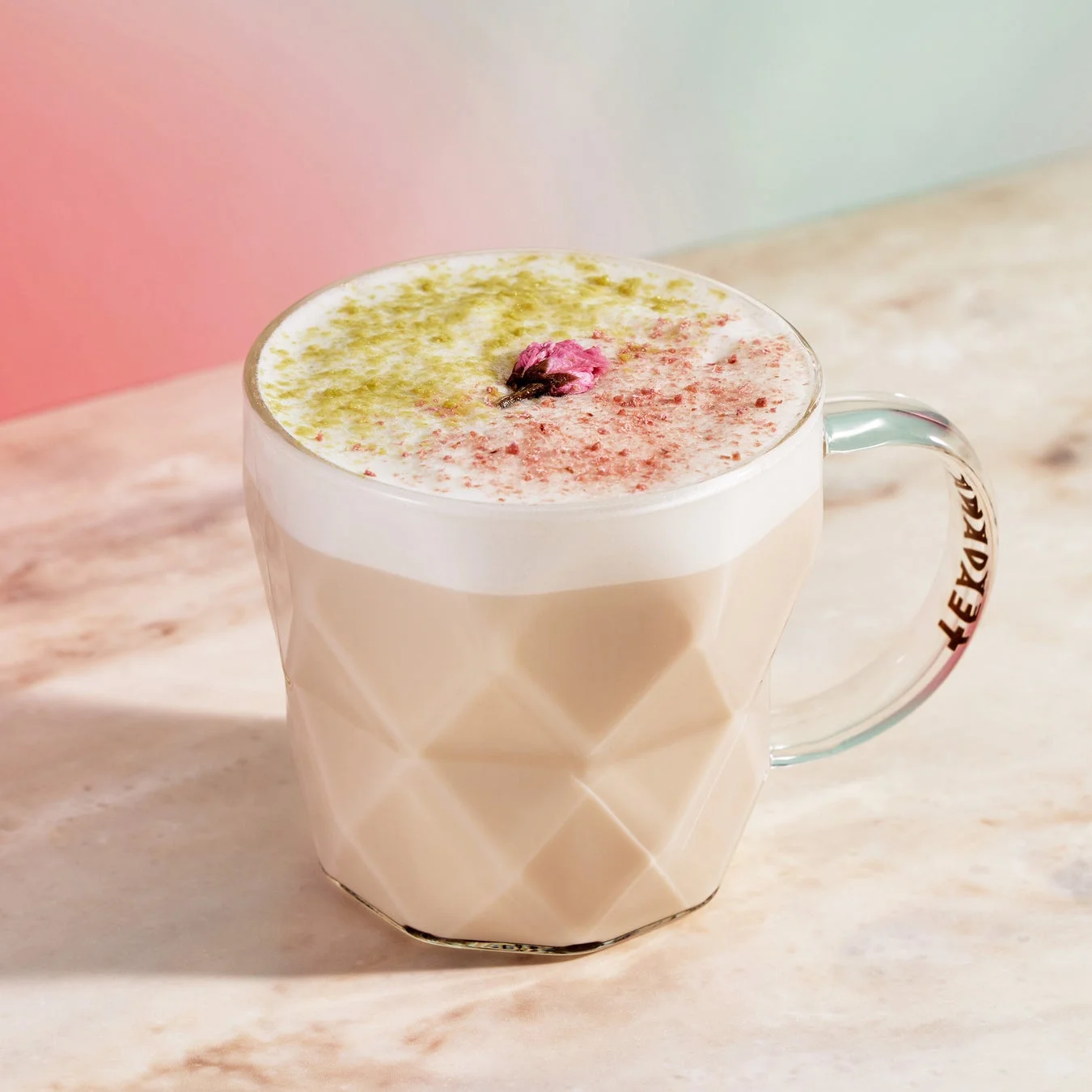 透明なファセット加工のマグカップに入ったラテで、クリーミーな泡の上に抹茶パウダーとピンクのパウダー、そして小さな桜の花が飾られています。背景は柔らかなパステルカラーで、全体的に美しく繊細な印象のドリンクです。
