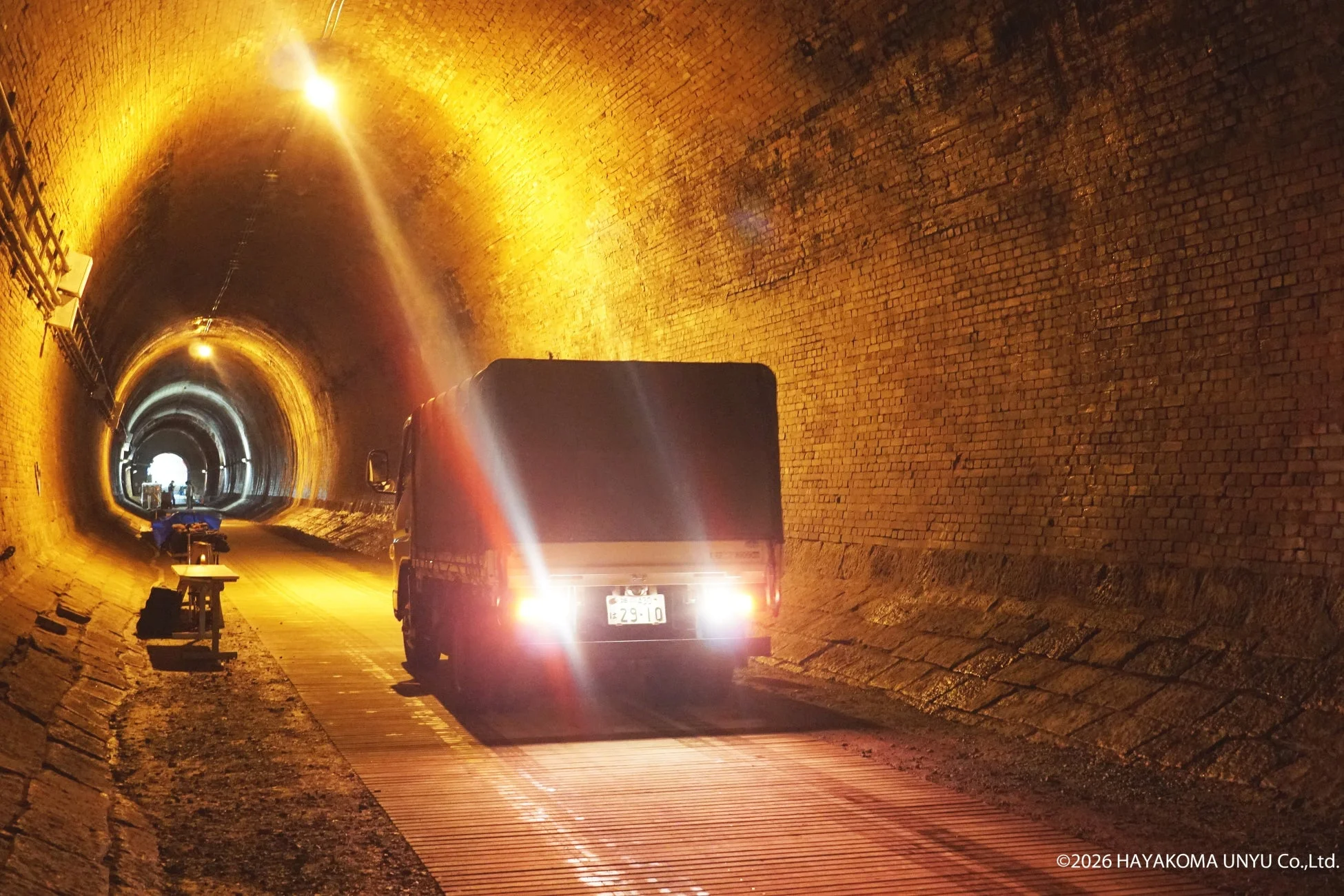 明るく照らされたレンガ造りの長いトンネル内をトラックが走行している様子を捉えた画像です。